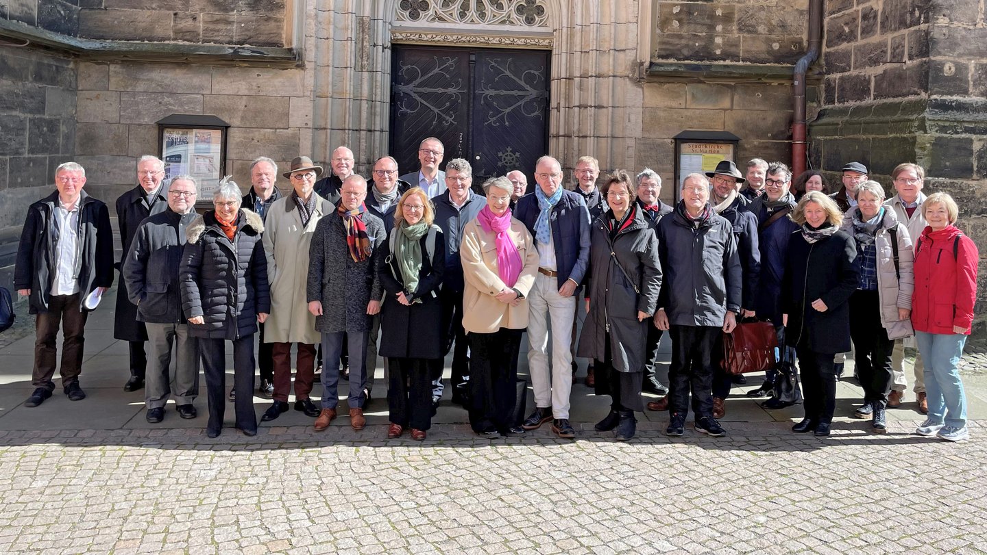 Eine Gruppe von Menschen steht im Sonnenschein vor einem Kirchenportal.